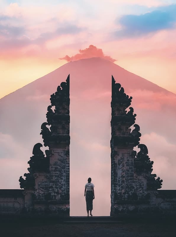 Žena vchádza do chrámu na Bali, ktorý má veľmi okázalý, architektonicky zaujímavý vchod, v pozadí vysoká hora.
