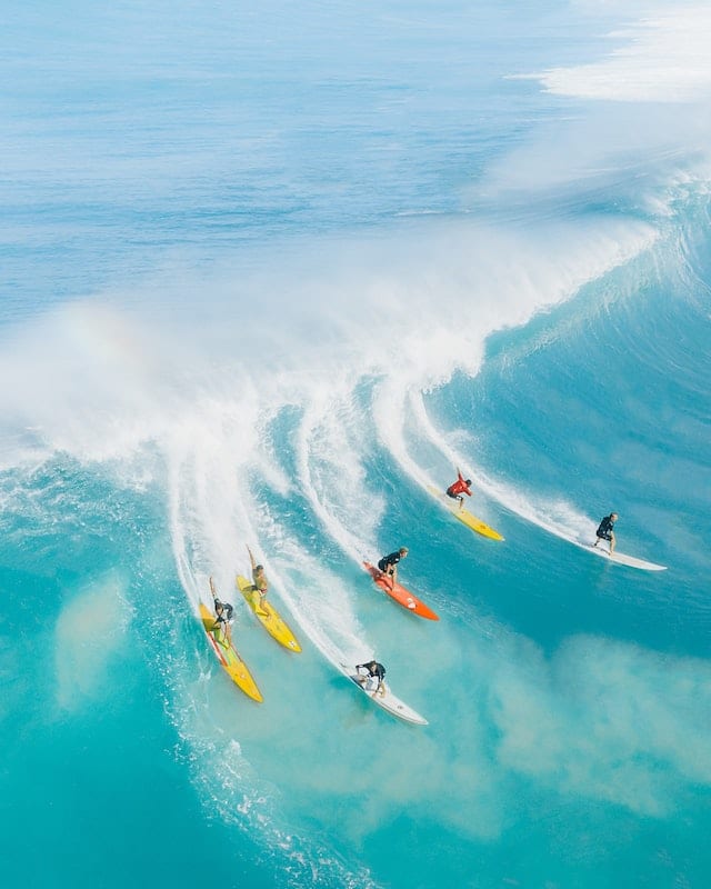 Priezračné modré more na Havaji so zdvíhajúcou sa vlnou, na nej sa schádza šesť surferov.